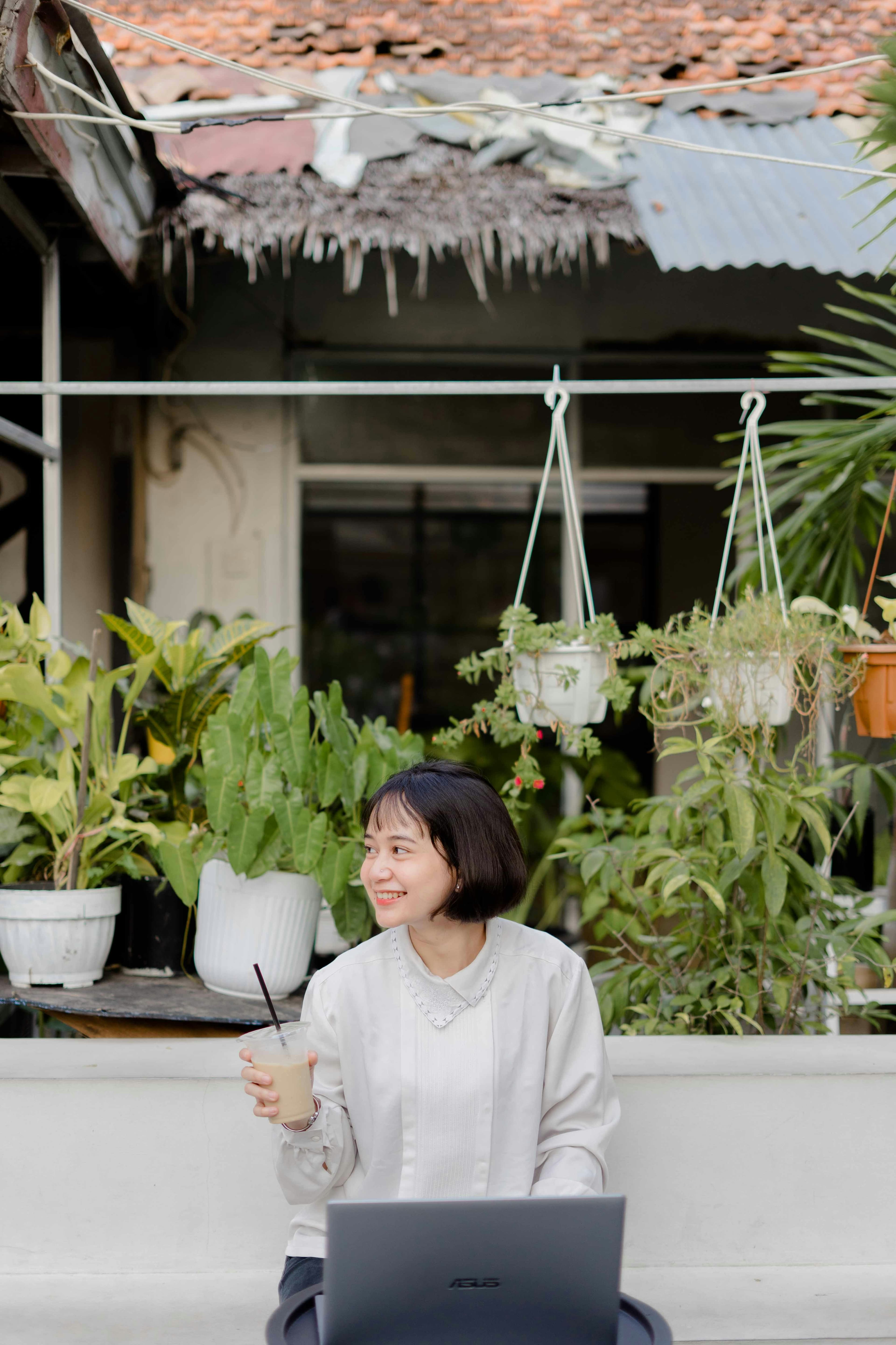 A young woman smiling outdoors with a laptop and an iced coffee.