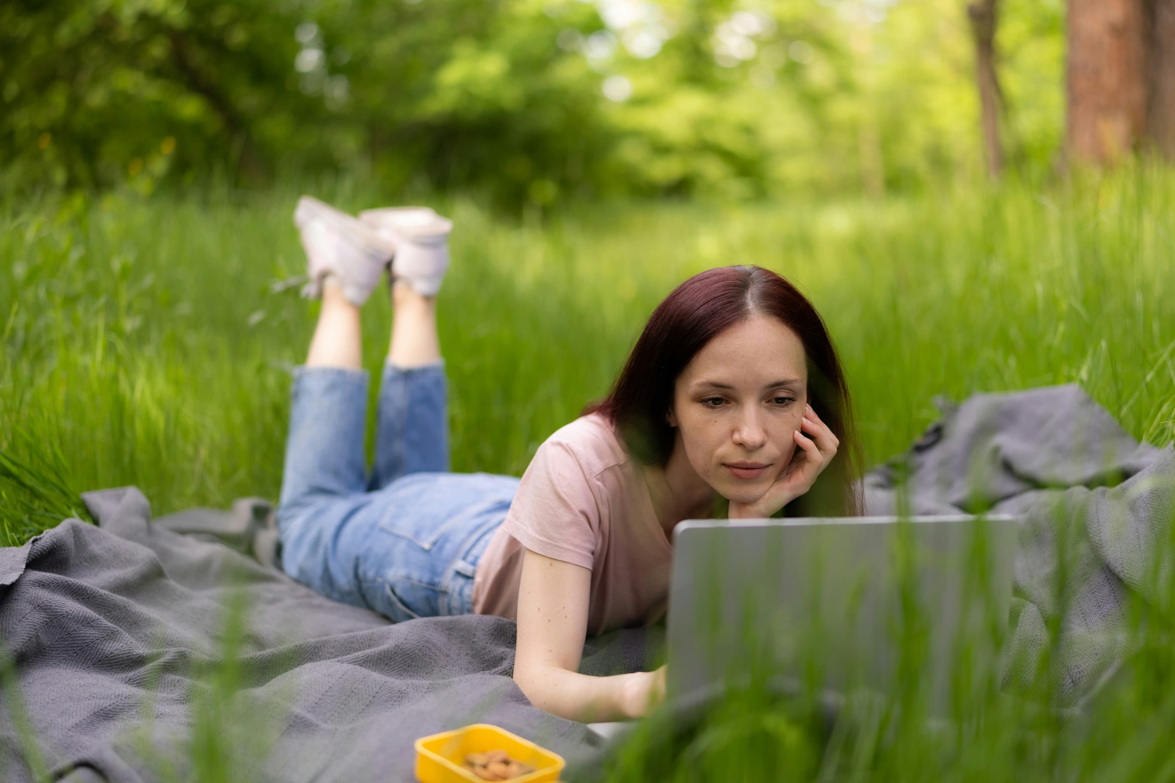 A woman lying on a blanket in the grass, reading closely on her laptop.