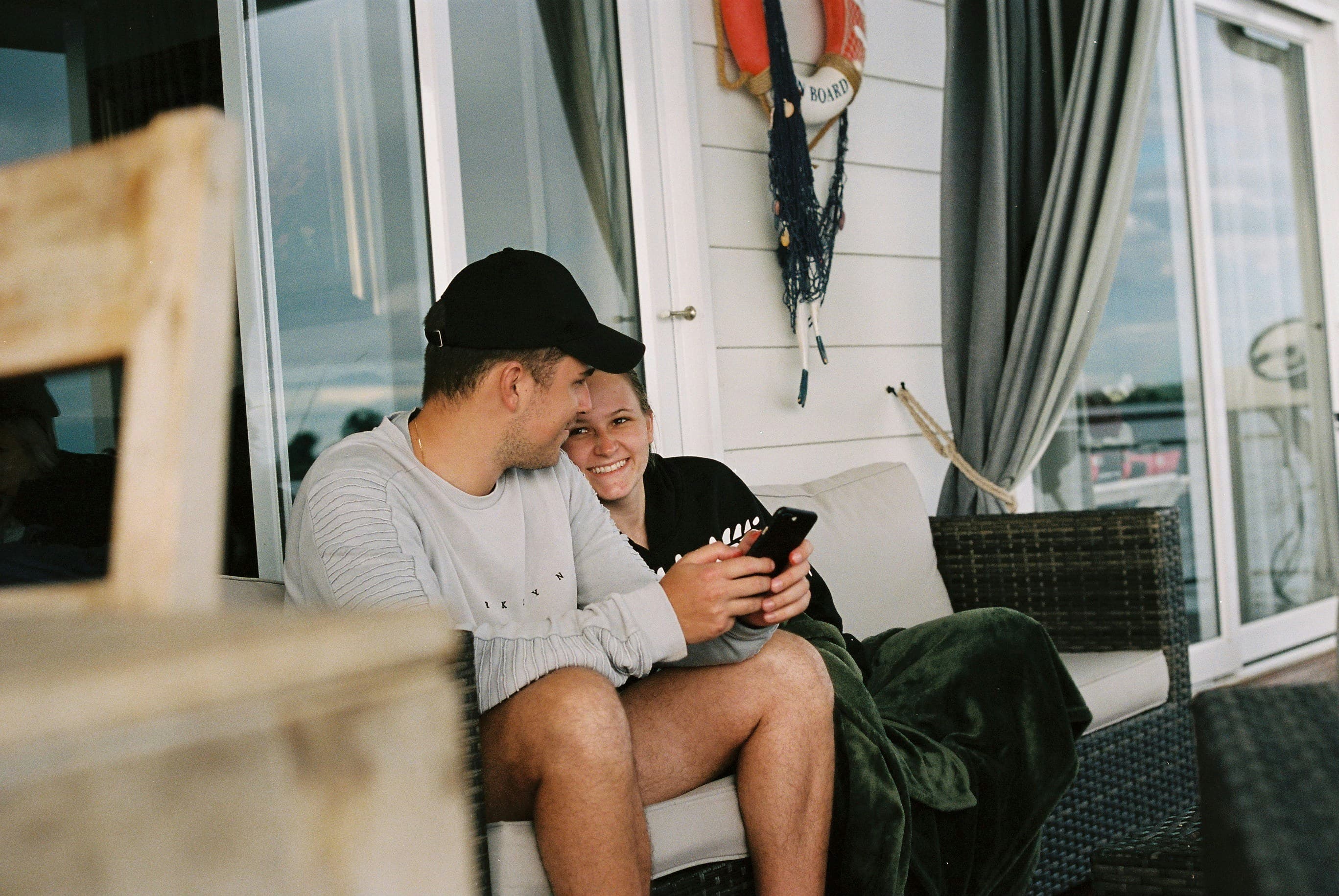 A young couple laughing over a phone on a waterside porch.