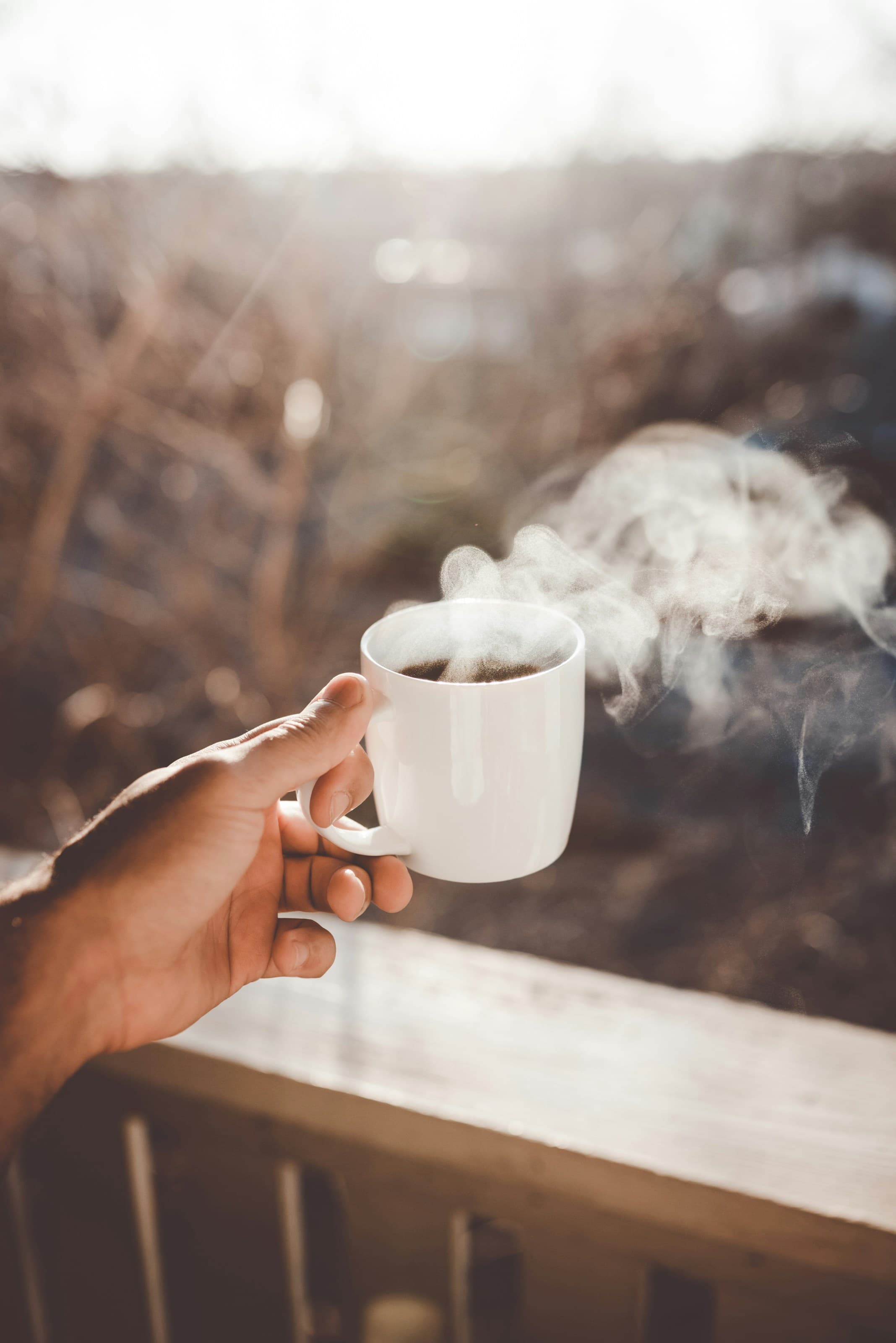 A hand holding a steaming mug of coffee on a porch in the morning sun.