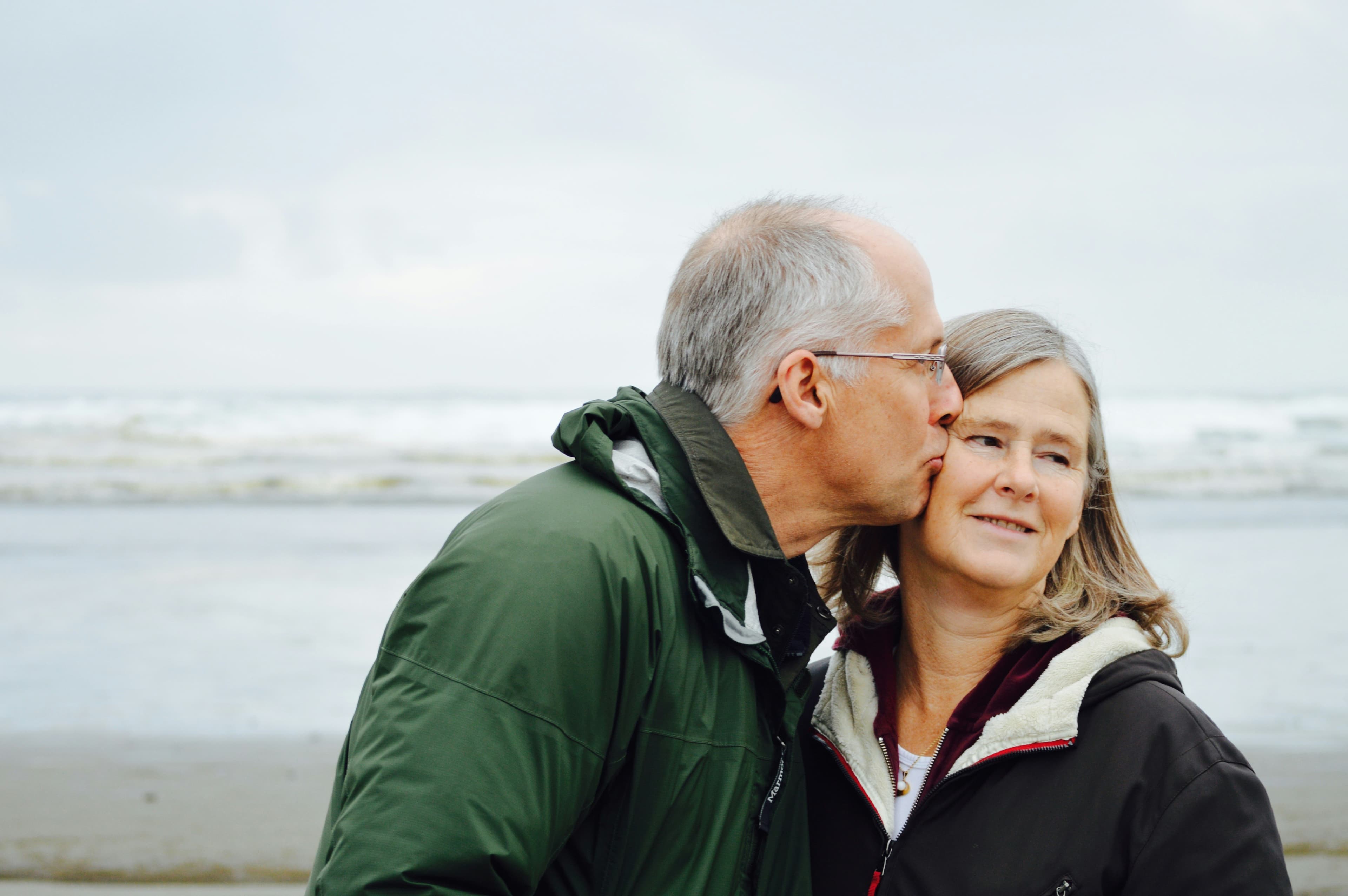 An older man kissing his wife on the cheek on a beach.