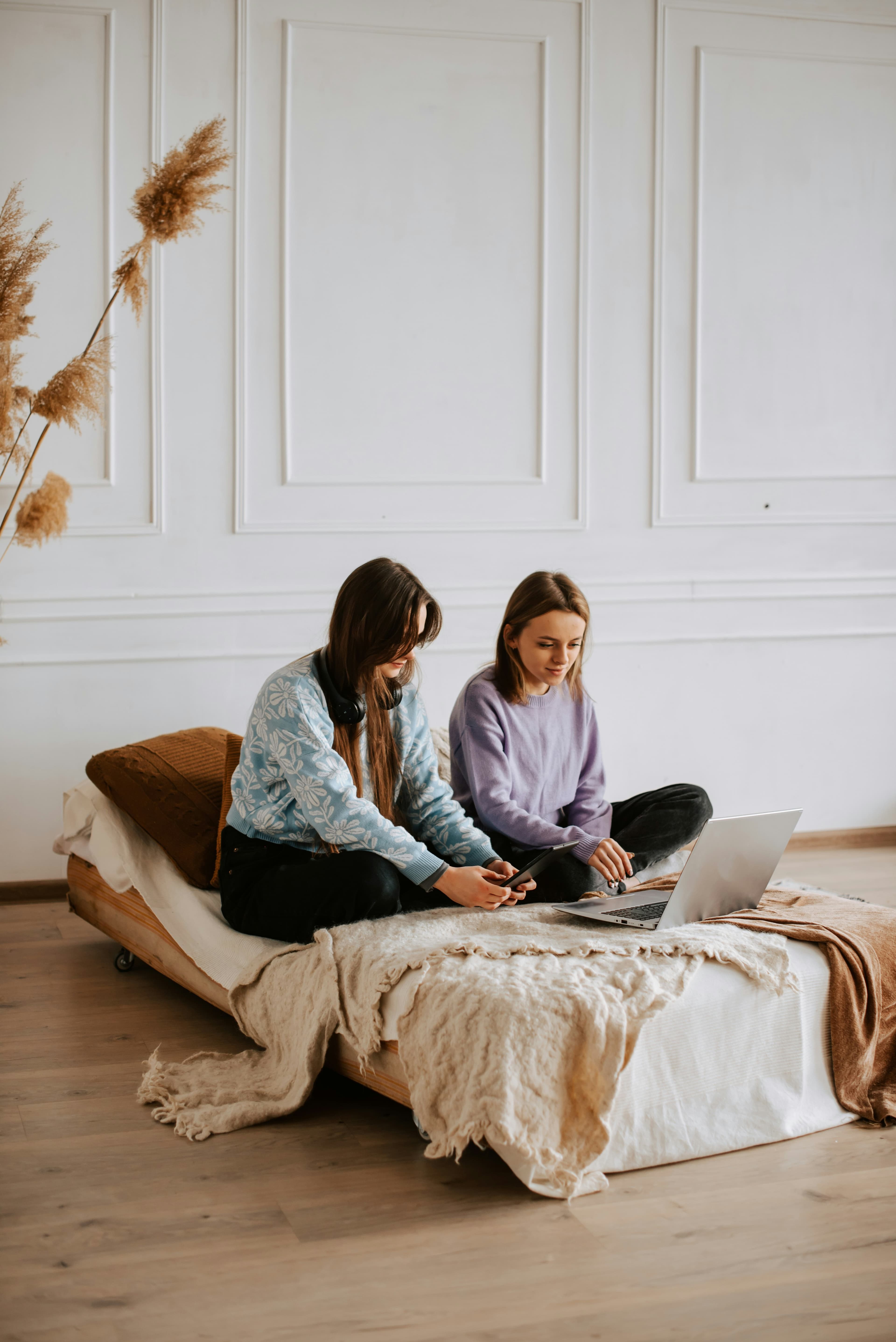 Two women sitting on a bed working through something on a laptop and a phone.