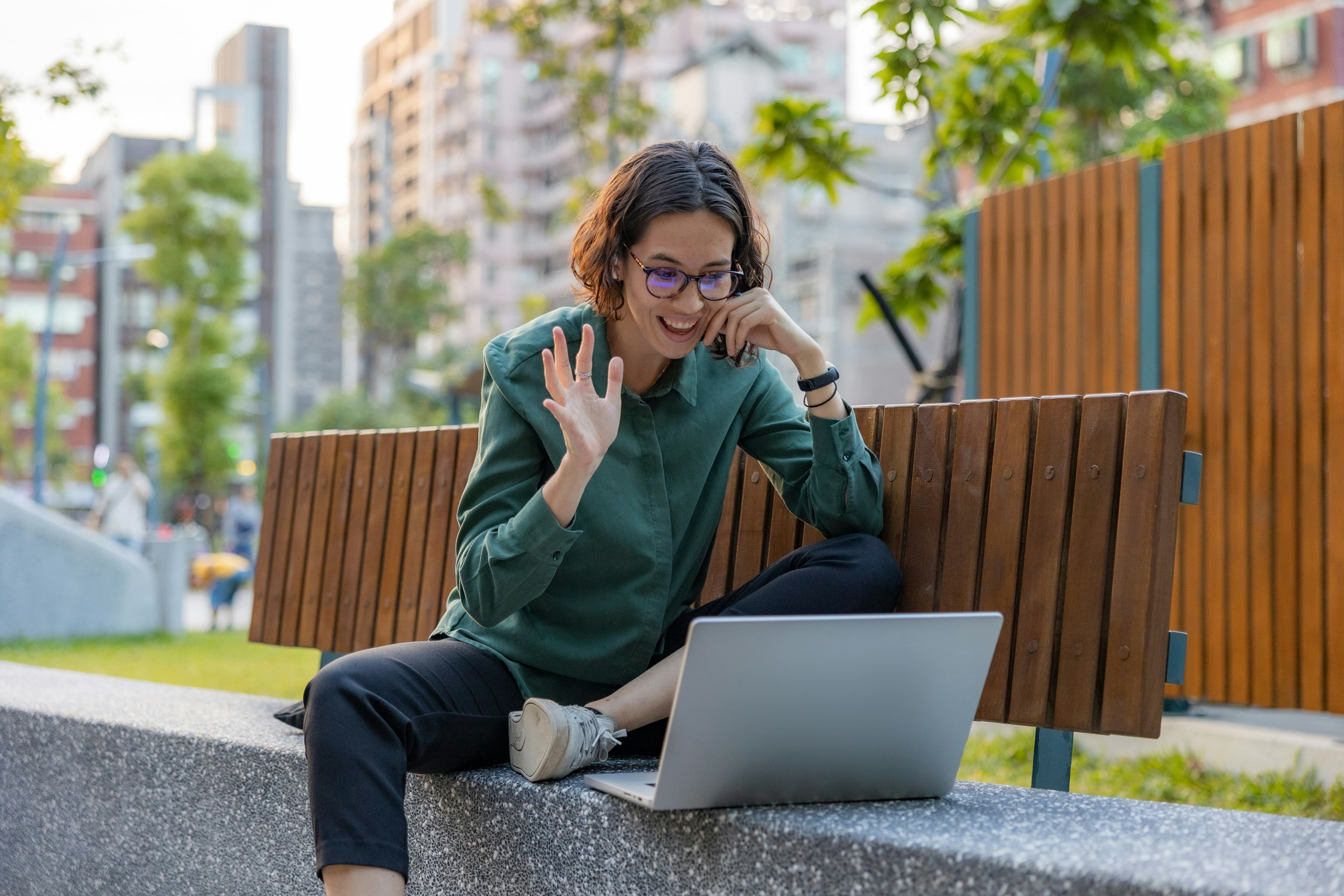 A woman waving to a video call from a laptop on a park bench.