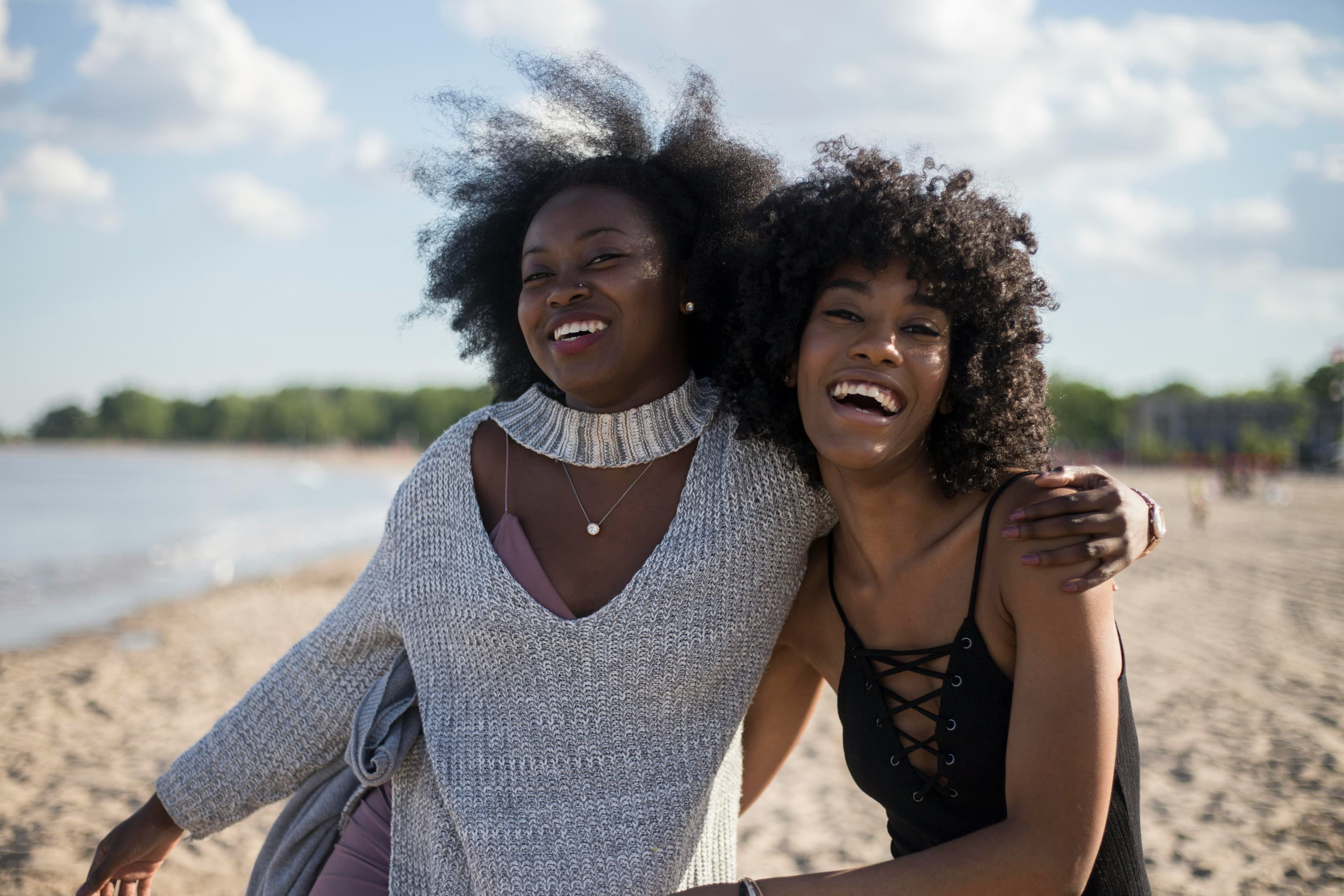 Two friends laughing arm in arm on a sunny beach.