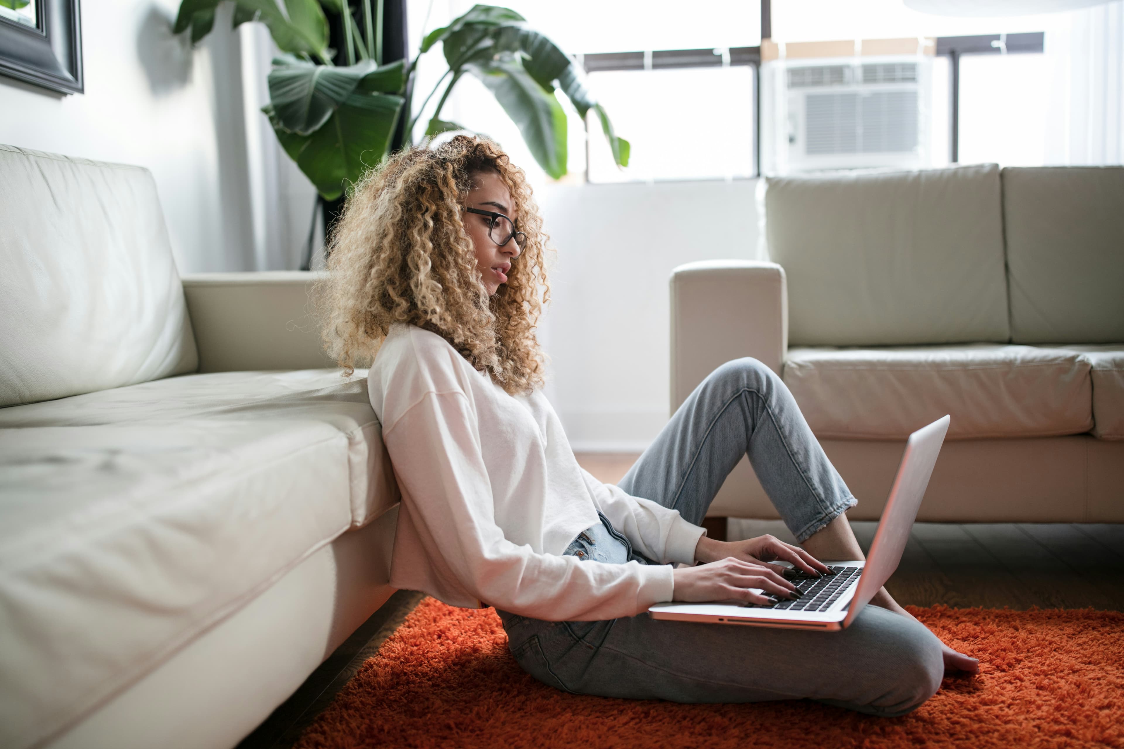 A woman sitting on a living room rug typing on a laptop.