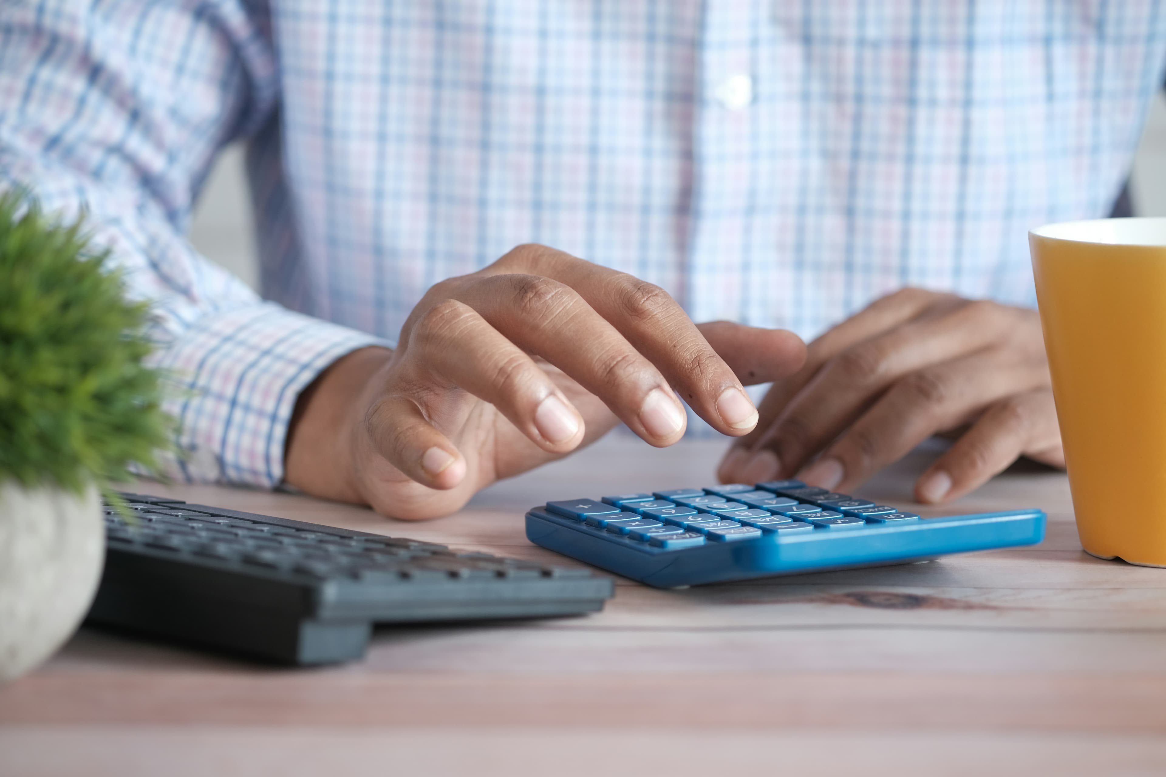Hands working a calculator at a desk beside a keyboard and a mug.