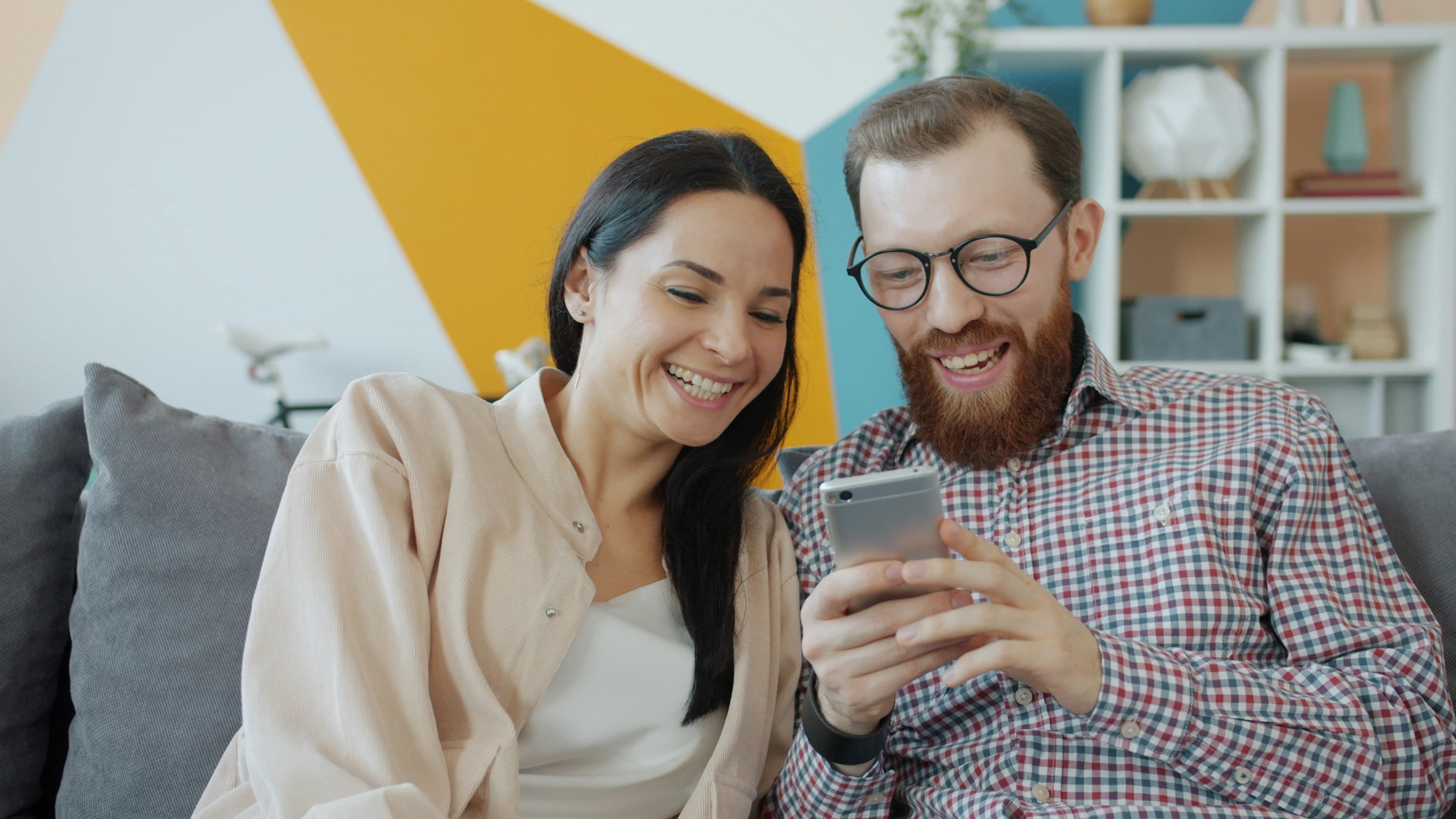 A couple on their sofa laughing at something on a phone.