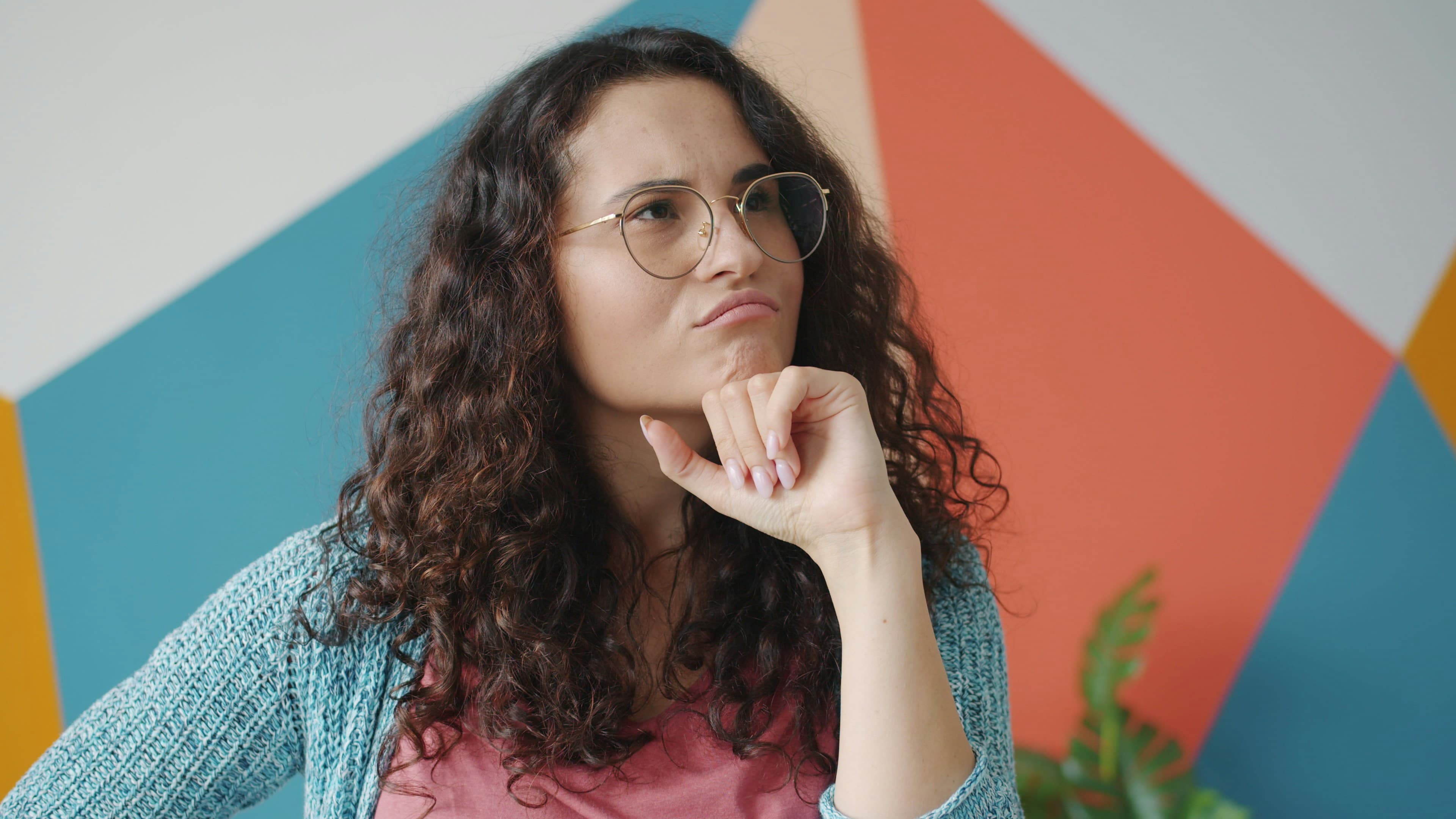 A woman in glasses weighing a decision, chin in hand.