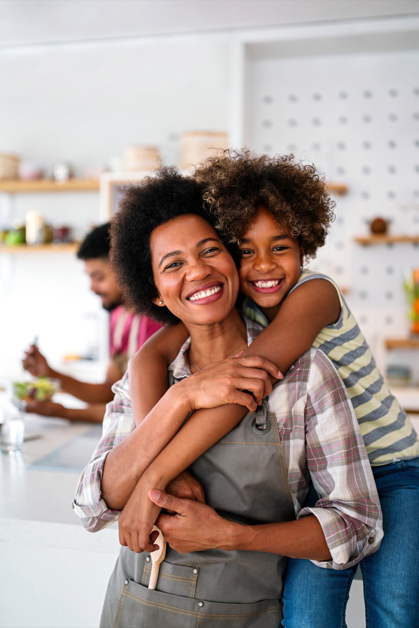 A mother and her young daughter laughing together in their kitchen, the rest of the family behind them.