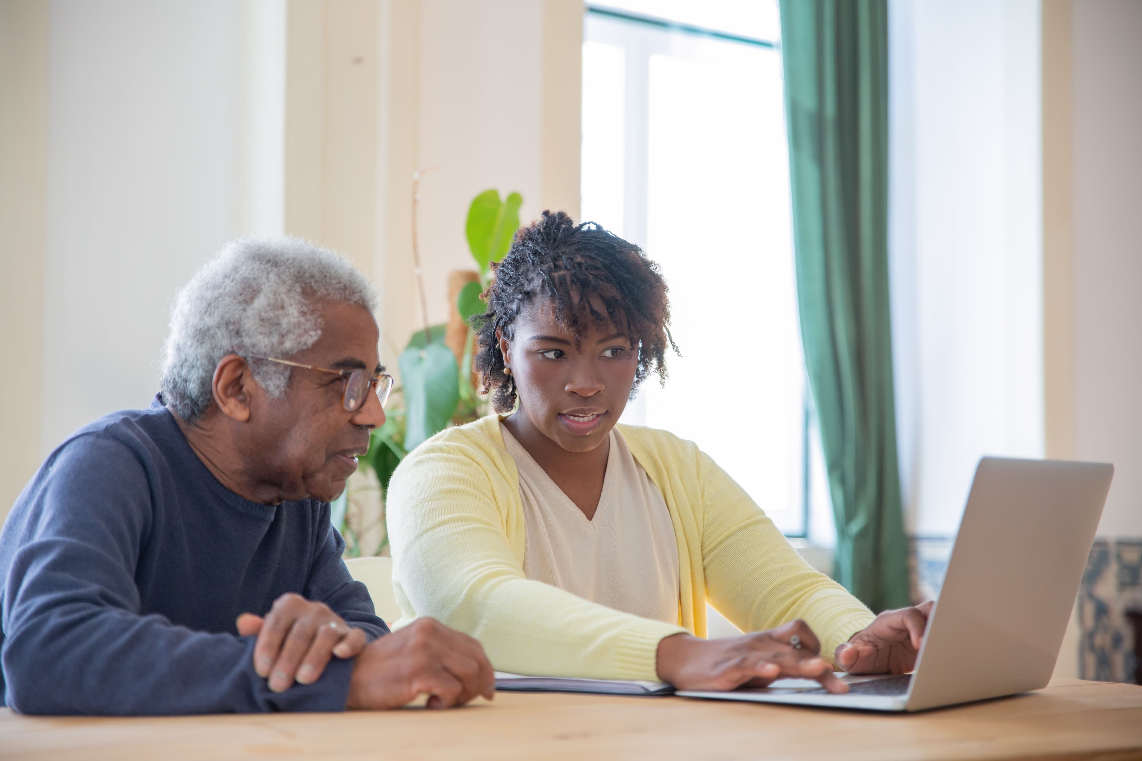 An older man and a younger woman sit together at a table, talking and looking at a laptop in a bright room.