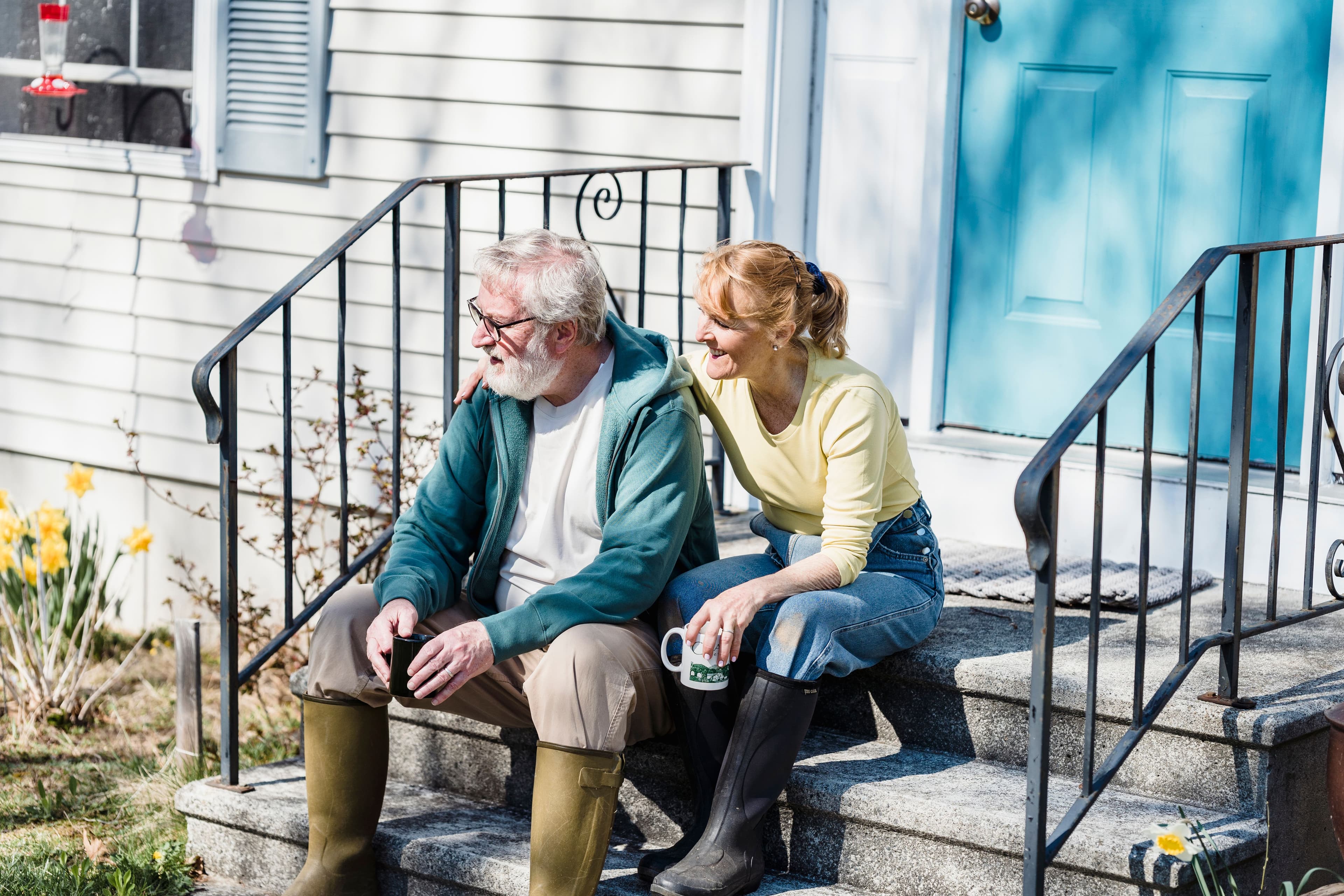 An older couple sits together on the front steps of their house, smiling and enjoying a sunny day.