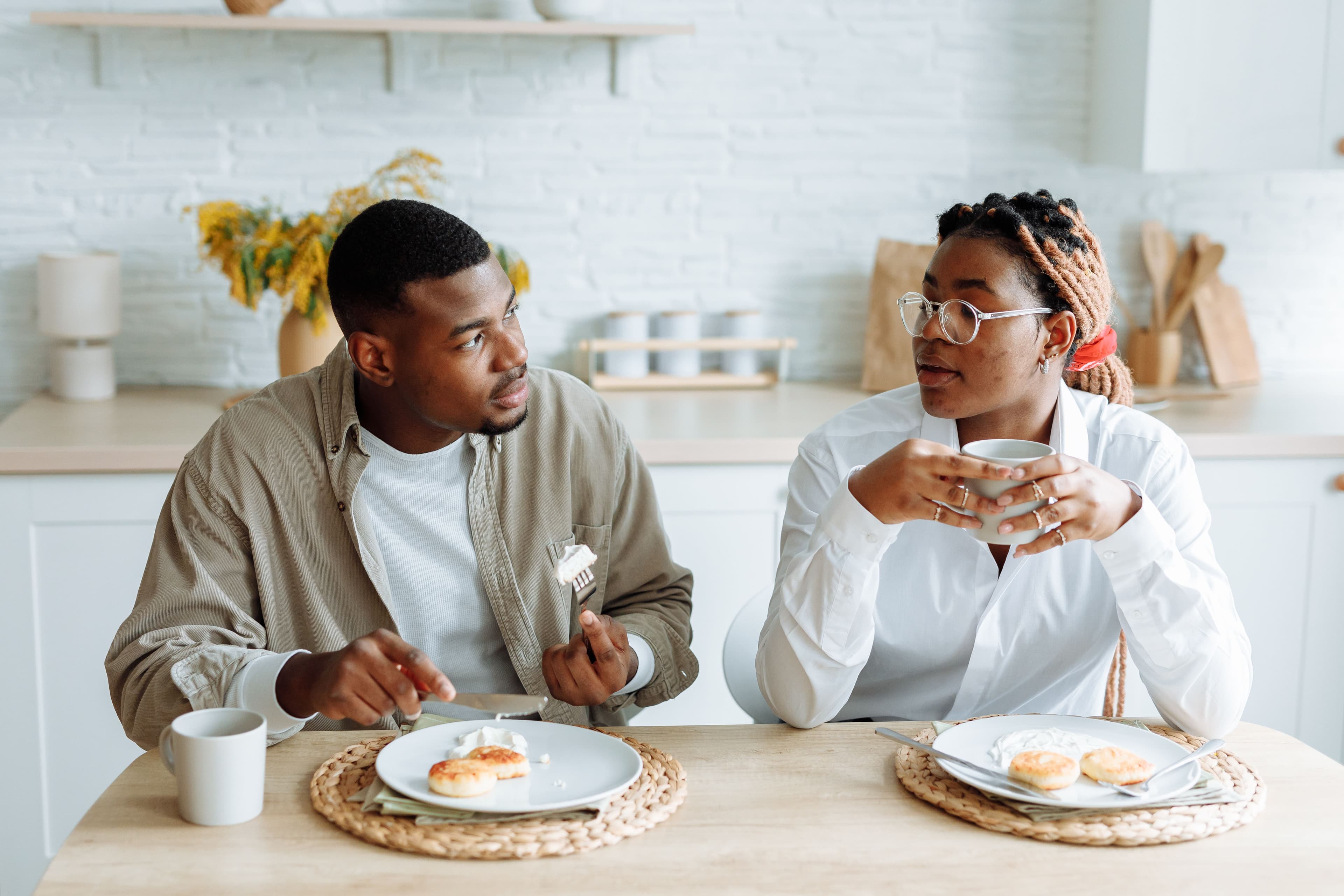 Two adults sit at a kitchen table having a calm conversation over coffee and pastries in a bright kitchen.