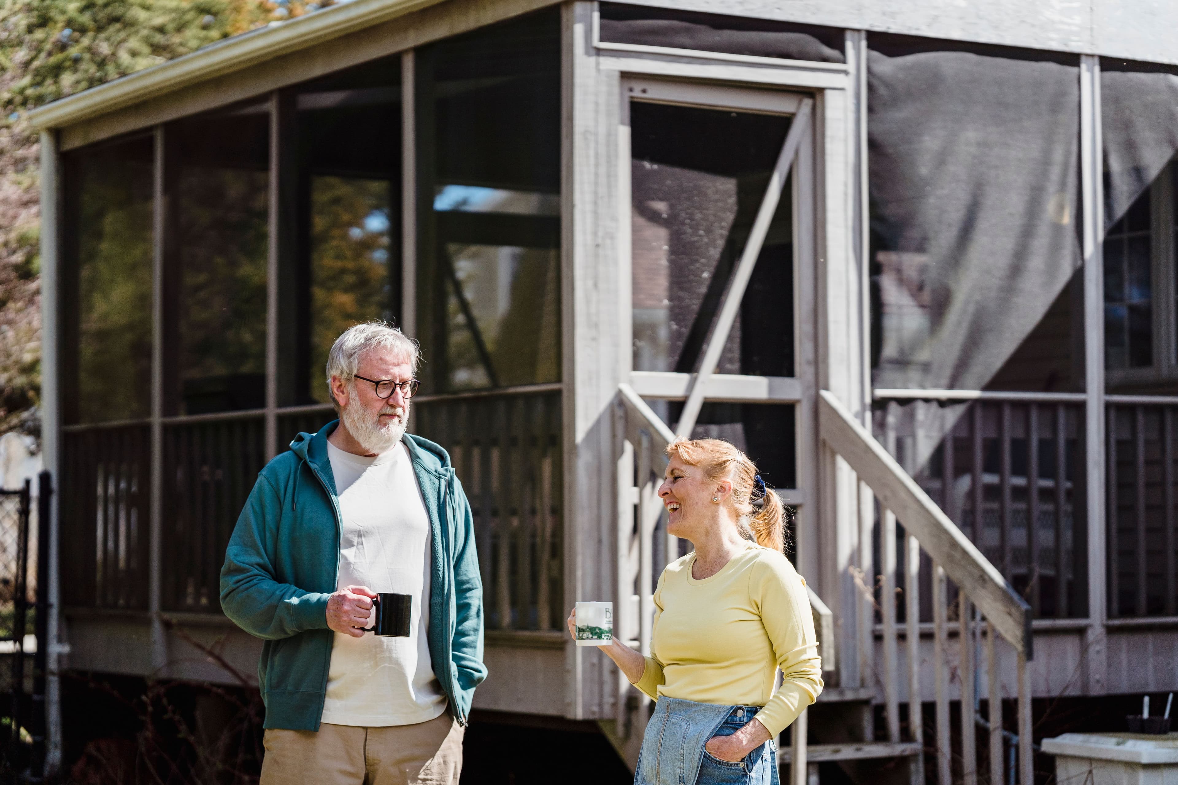 An older man and a woman stand outside a porch, holding mugs and having a calm conversation in the sunlight.