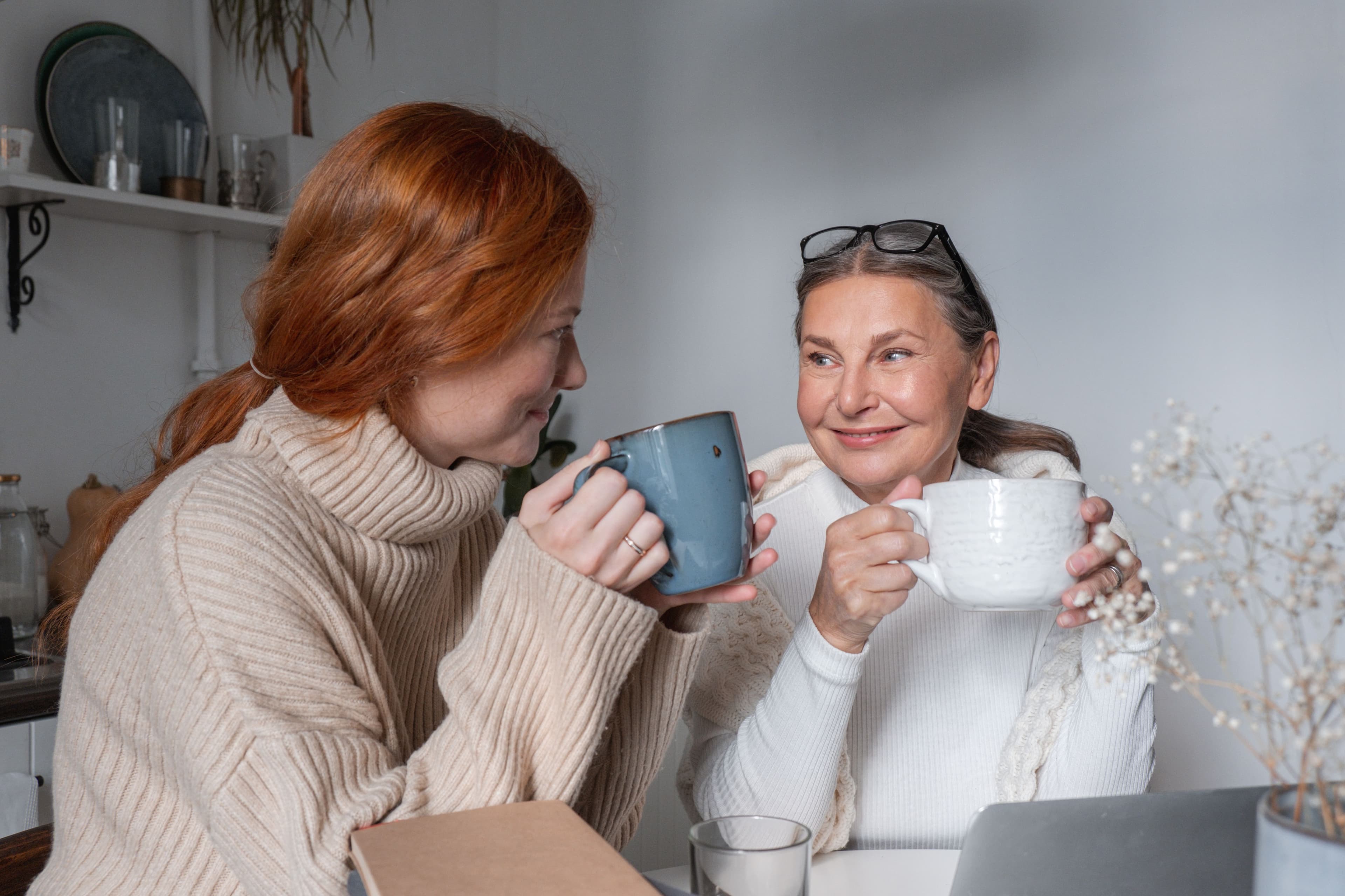 An older woman and a younger woman sit at a kitchen table smiling and talking while holding mugs of coffee or tea.