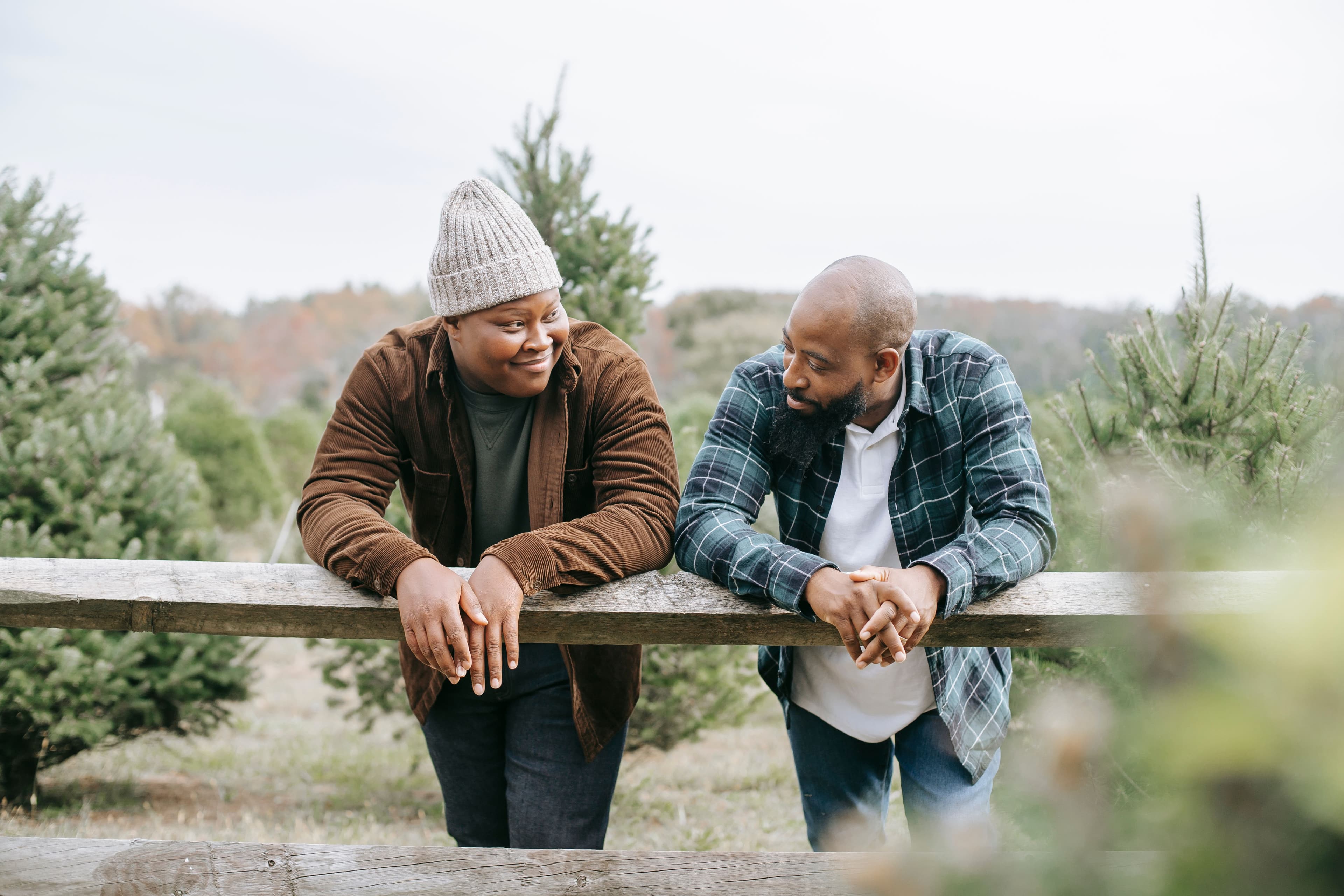 Two adults lean on a wooden fence outdoors, talking and smiling at each other in a calm and supportive way.