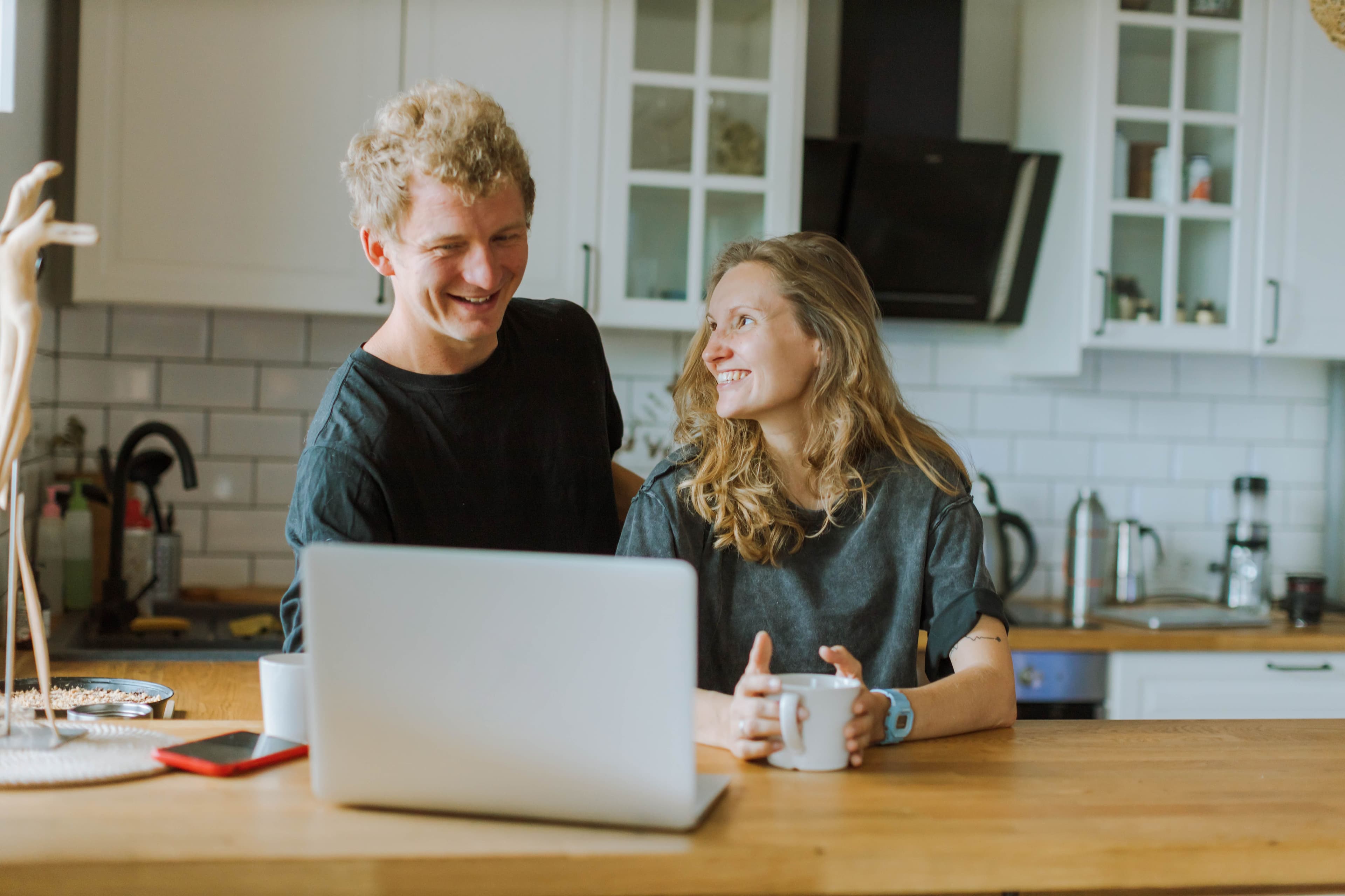 A smiling couple sits at a kitchen table with a laptop and coffee mugs, looking relaxed and happy together.