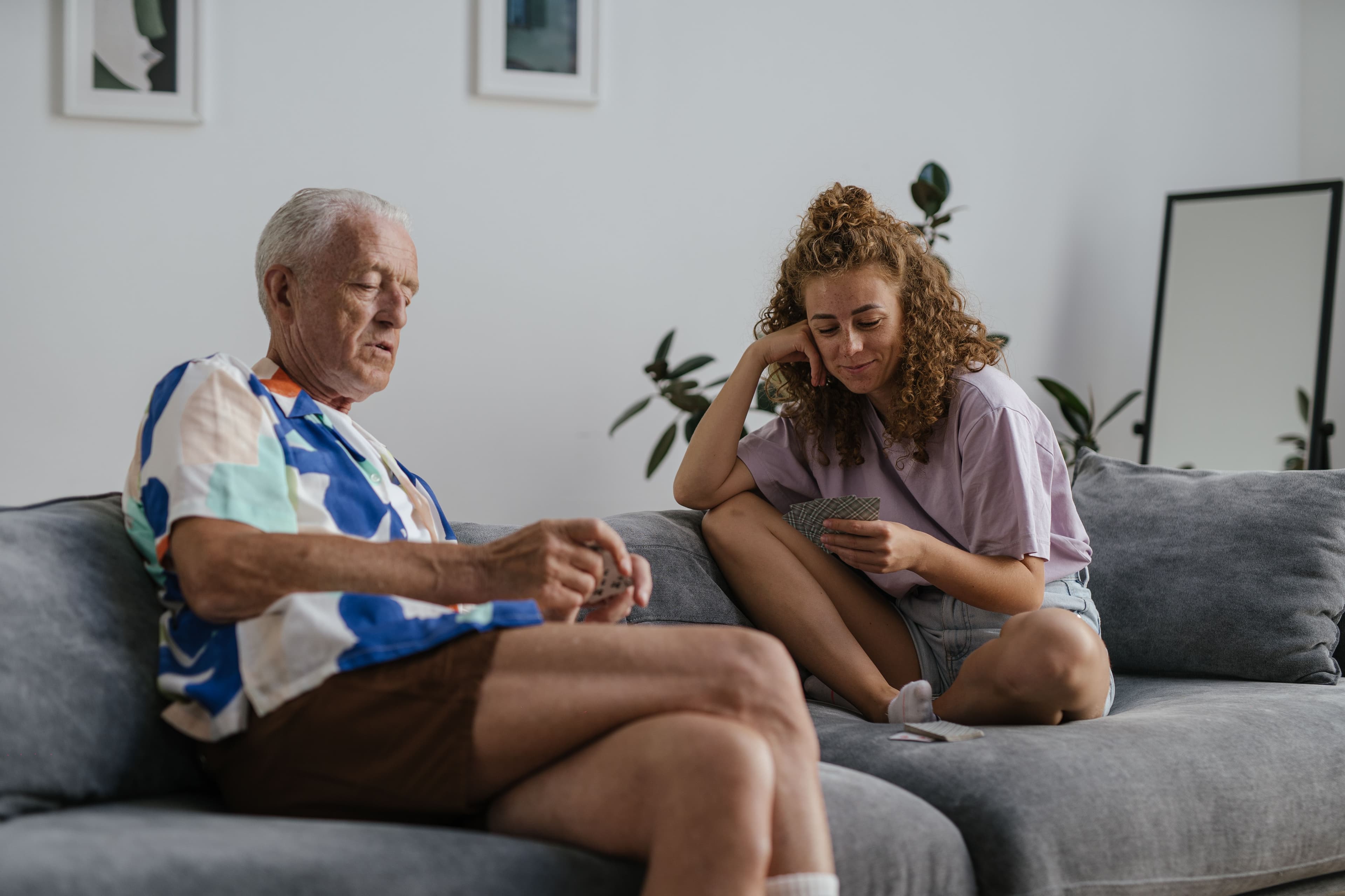 An older man and a younger woman sit together on a couch playing cards and enjoying a calm moment at home.