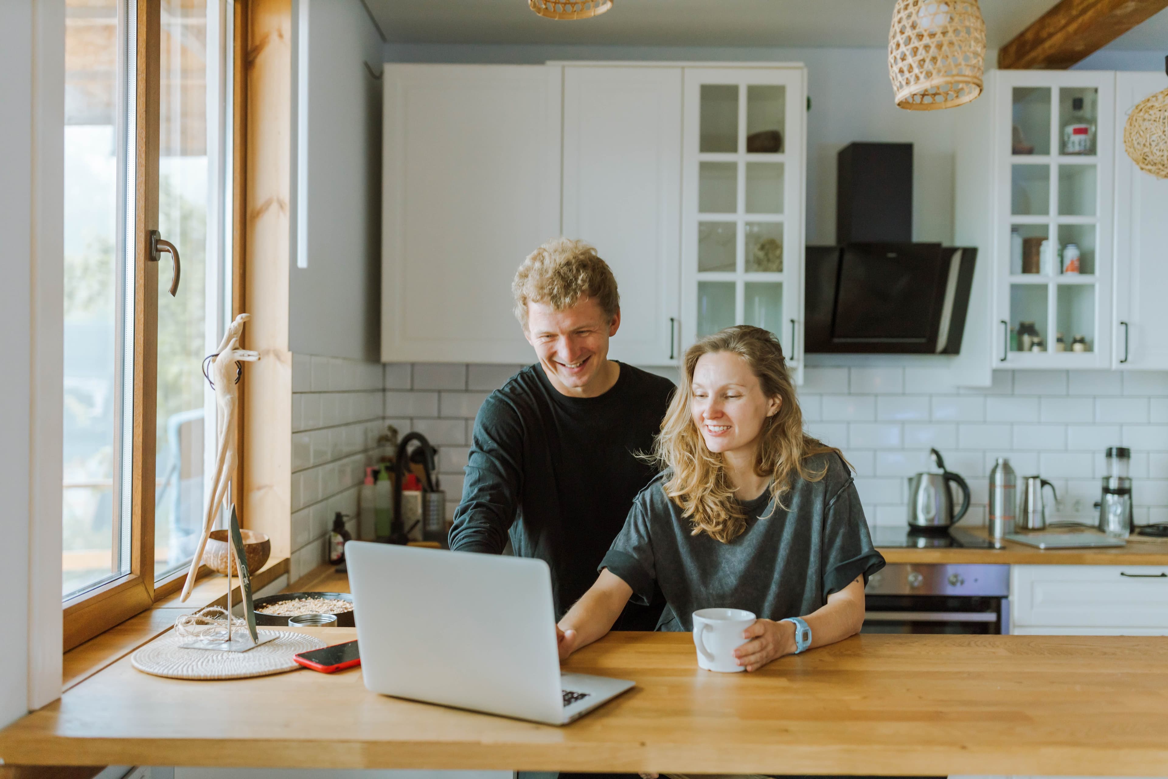A smiling couple sits at a kitchen table with a laptop, one holding a mug, both appearing relaxed and happy.