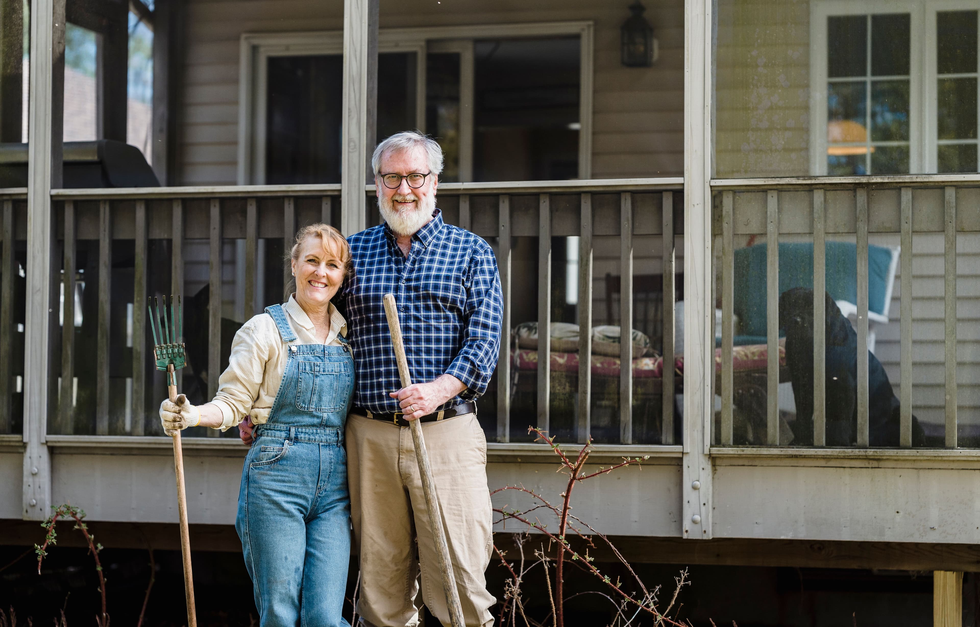 A smiling couple stands together in front of their house holding gardening tools, with a dog visible on the porch behind them.